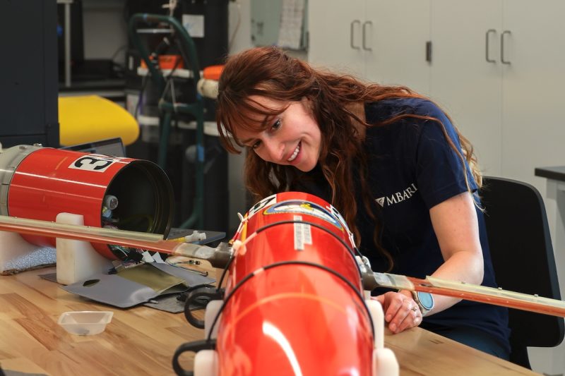 A scientist examines an oceanographic robot on the wooden workbench of an engineering laboratory. She is dismantling the cylindrical orange plastic housing and examining the electronics inside. In the background are white cabinets, black shelves, and various pieces of engineering equipment.
