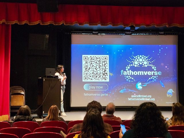A scientist stands on the left side of an auditorium stage behind a black podium with a laptop on it. She is standing on a stage framed with red curtains, next to a projector screen displaying a QR code and the Fathomverse project logo. In the foreground are theater seats occupied by attendees.