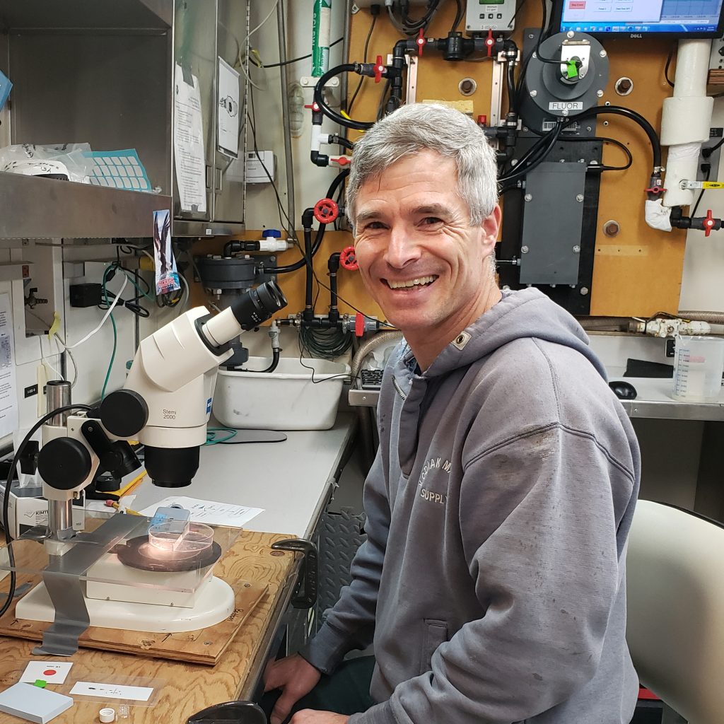 edited image of Rob Suryan sitting at a lab bench in front of a microscope. He is looking at the camera