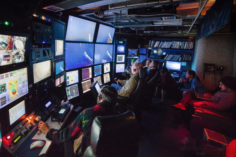 A team of six researchers sit in the darkened control room of an MBARI research vessel while watching a wall of video monitors streaming live video and data from a robotic submersible deployed in the deep sea. In the center, a submersible pilot wearing an olive shirt and a headset sits in a black leather chair and controls the robot with a black joystick. To the left is a second pilot in a red-and-green flannel shirt and headset observing screens displaying data from the ship and submersible.